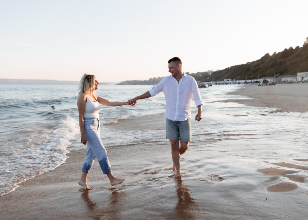 A couple walks barefoot along the shoreline, holding hands and smiling at each other, with waves gently touching their feet and hills in the background.