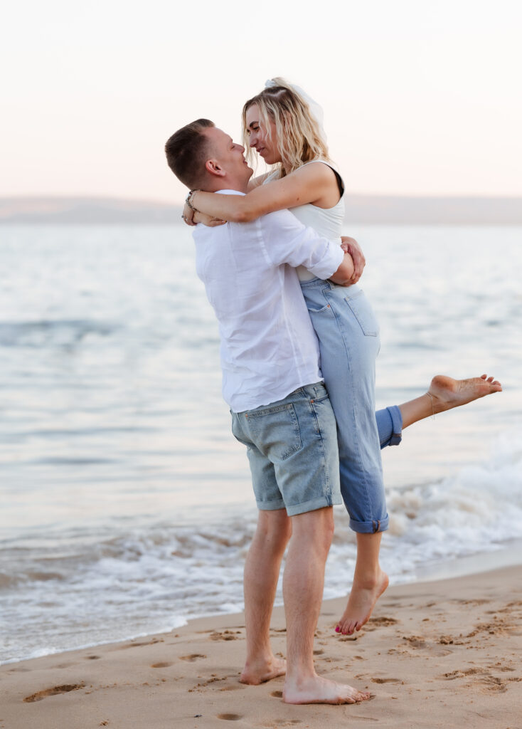 A man lifts a woman in his arms on a sandy beach near the water. They smile at each other, and she has one leg raised. The setting is bright and calm, with gentle waves in the background.