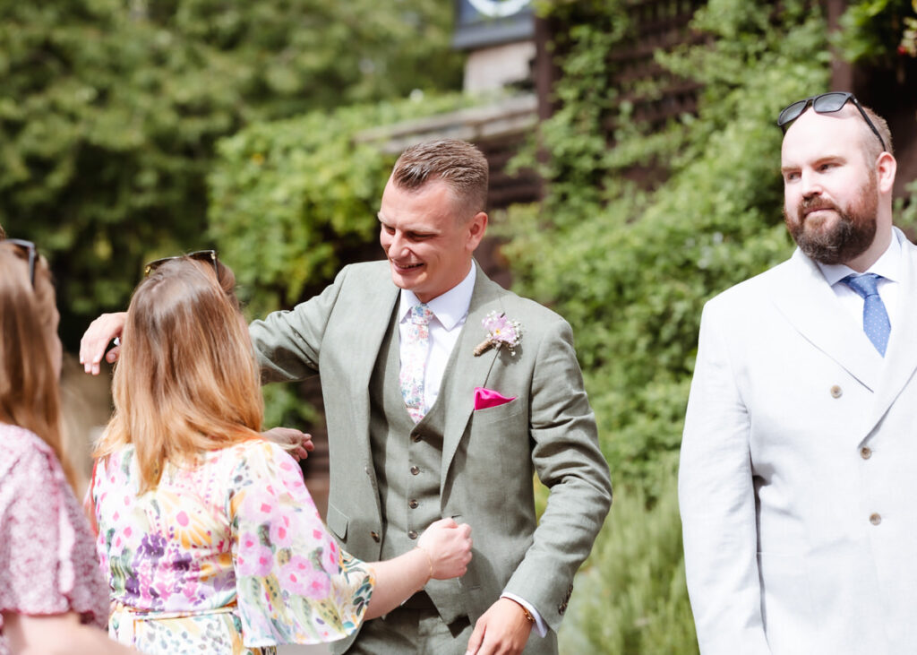 A man in a green suit smiles while greeting a woman in a floral dress outdoors, with another man in a white suit and blue tie standing nearby. Lush greenery is in the background.
