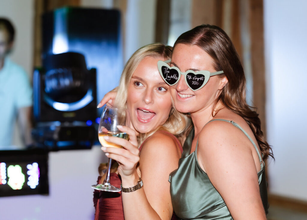 Two women smiling at a party, one holding a glass of wine and the other wearing novelty sunglasses that read The Smiths and The Augusts 2023. Both appear to be having fun.