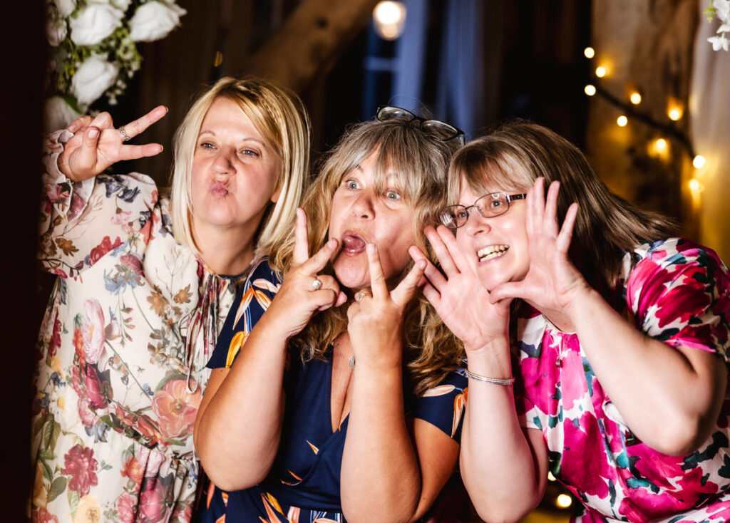 Three women posing playfully for a photo, making funny faces and hand gestures. They are wearing colorful floral dresses and standing close together with decorative lights and flowers in the background.