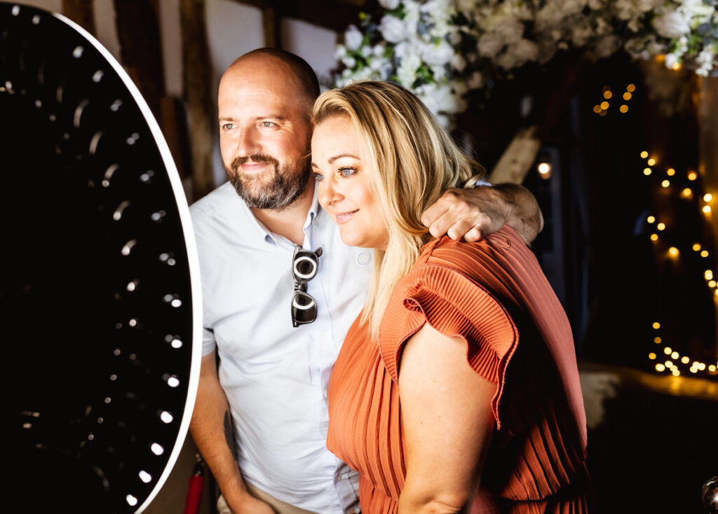 A man and woman pose closely together, smiling at a photo booth with bright ring lighting. The background features floral decorations and string lights, creating a festive atmosphere.