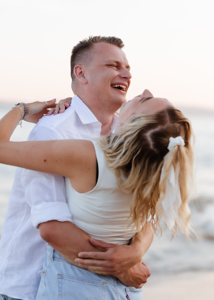 A couple laughs and embraces on a beach, with the person in front smiling up at the other. The background is blurred with soft colors, suggesting sunset by the sea.