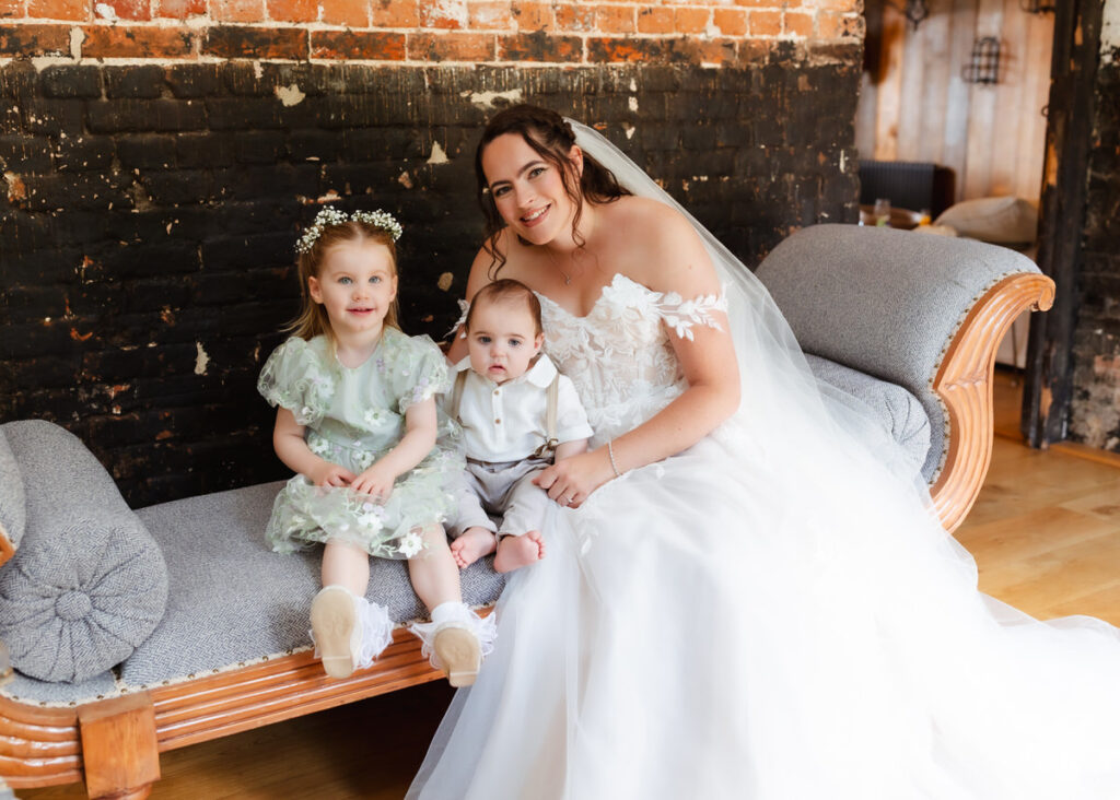 A bride in a white dress sits on a couch with two young children, a girl in a green dress and a baby boy in light clothes, smiling in front of a rustic brick wall.