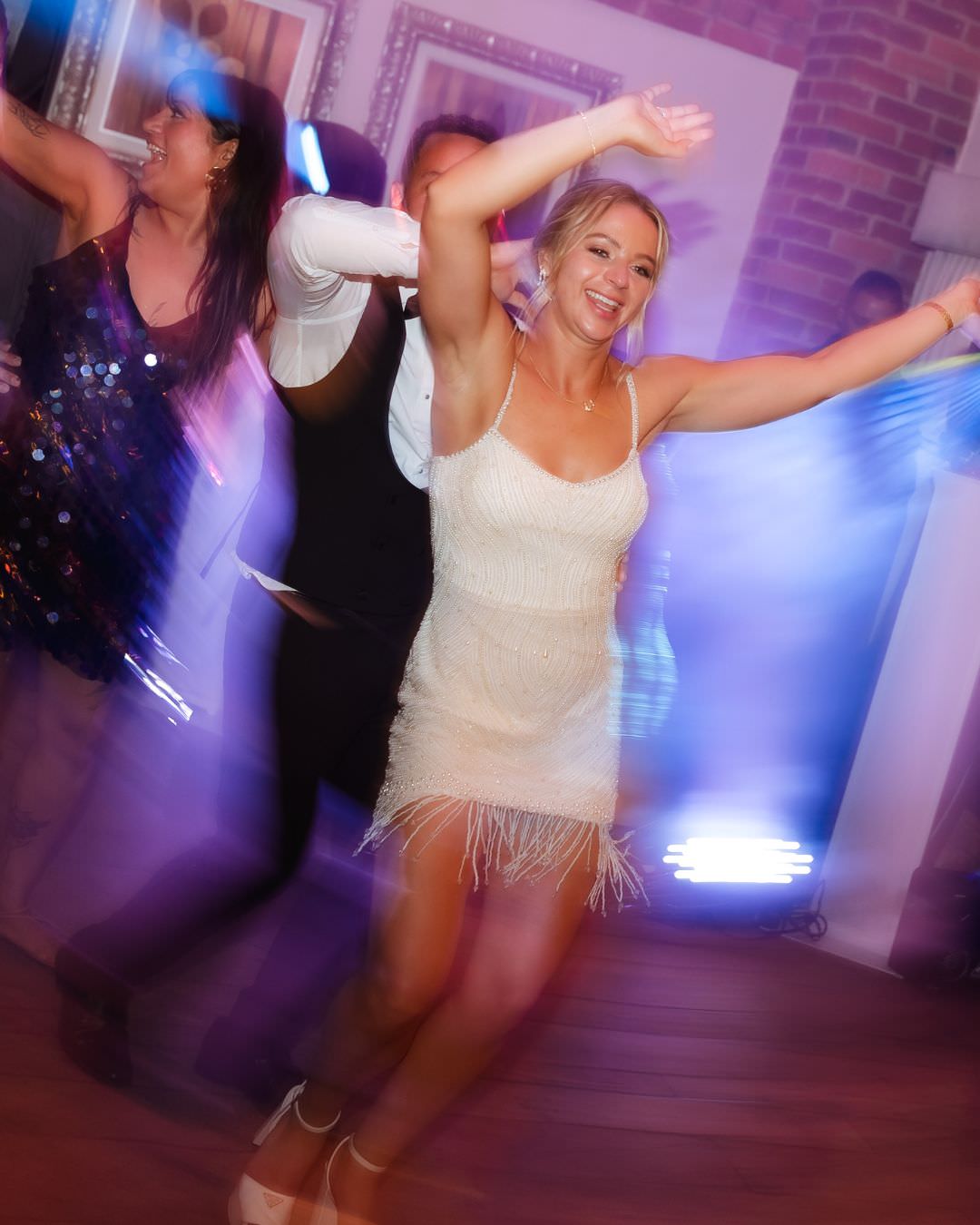A bride in a white beaded fringe dress dances with guests at a country-house wedding, captured with motion blur and atmospheric blue lighting.