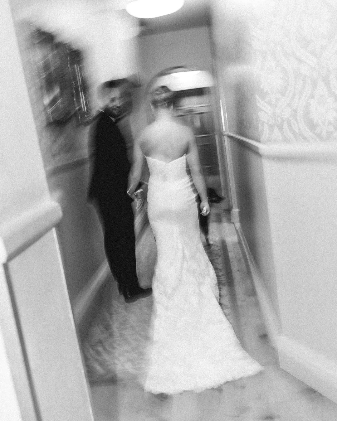 A bride in a strapless gown and a man in a tuxedo walk down a hallway together in a motion-blurred black and white photo by a country-house wedding photographer.